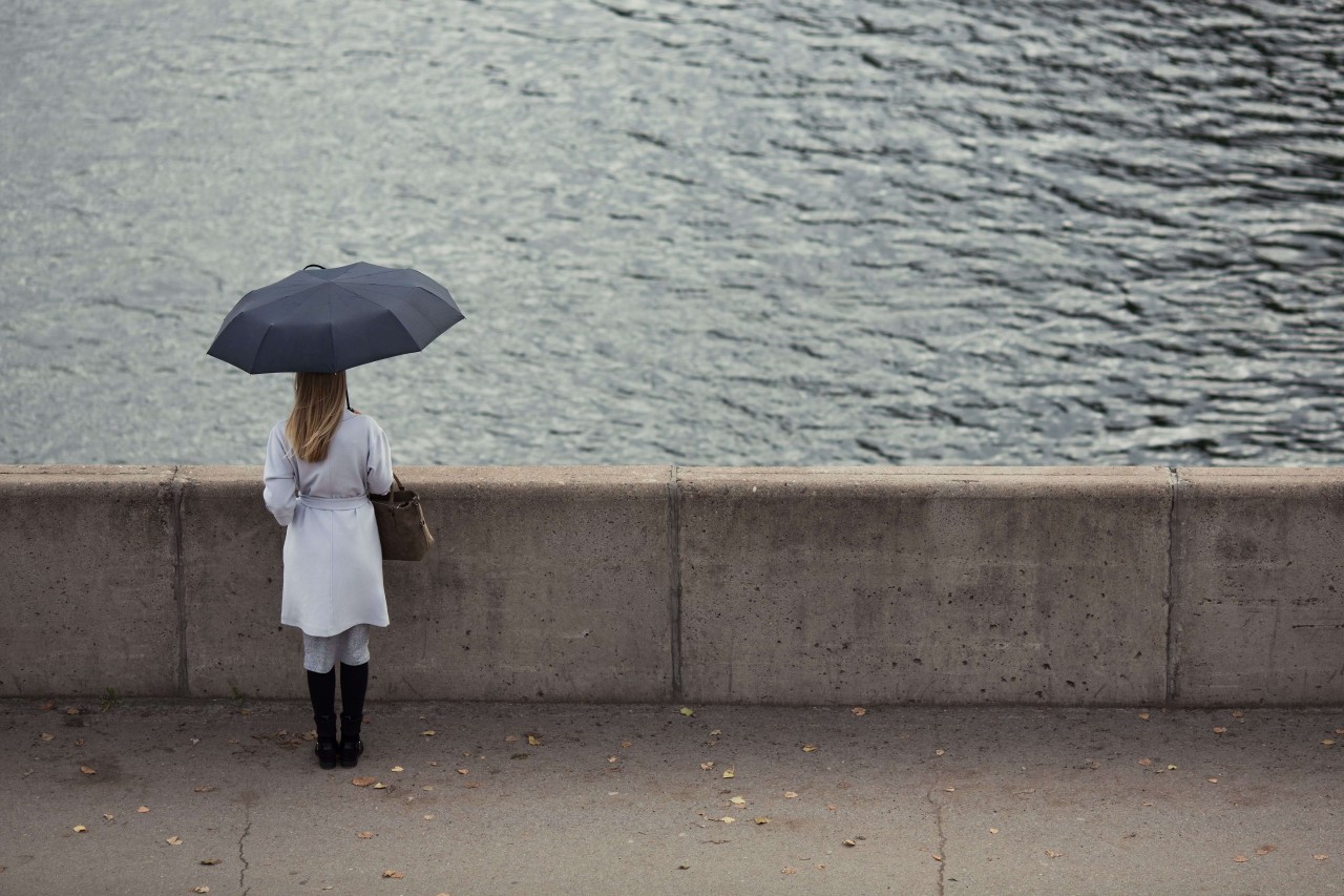 Nainen seisoo yksin ja katsoo kaukaisuuteen sateisessa säässä sateenvarjon alla